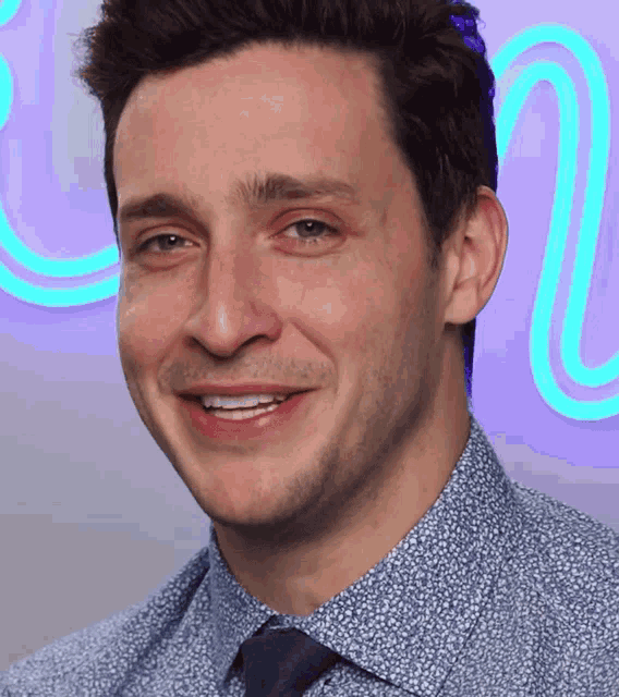 a man wearing a blue shirt and tie smiles in front of a neon sign