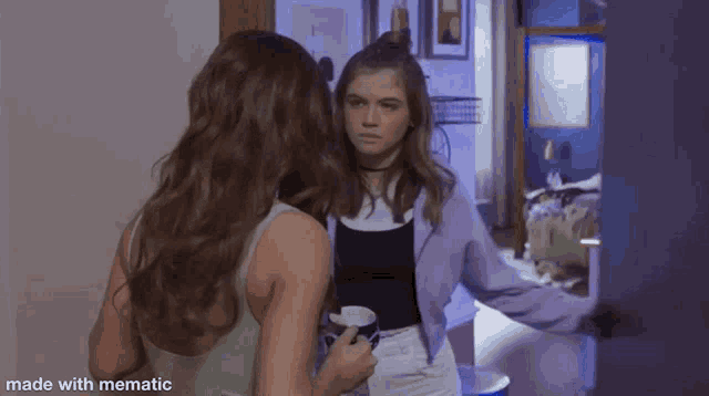 two girls are standing next to each other in a room . one of the girls is holding a cup of coffee .