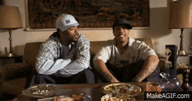two men are sitting on a couch with one wearing a hat that says ' ii ' on it