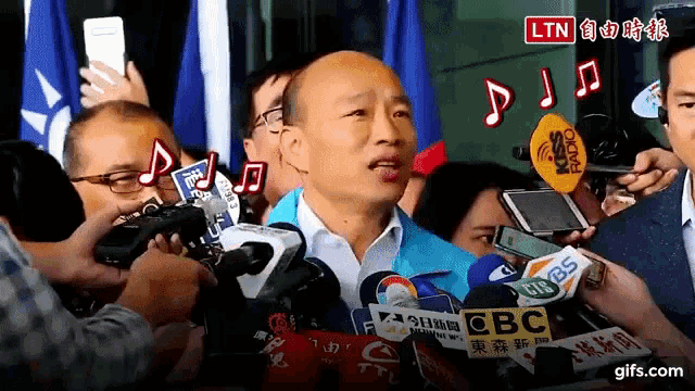 a man in a blue jacket is surrounded by many microphones including one that says cbc