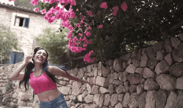 a woman in a pink crop top stands in front of a stone wall with pink flowers
