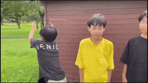 three young boys are standing in front of a wooden building and one of them is wearing a yellow t-shirt that says venic