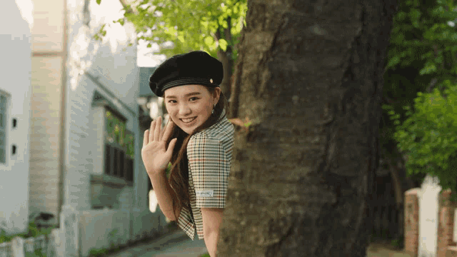 a woman wearing a plaid shirt and a black beret waves from behind a tree