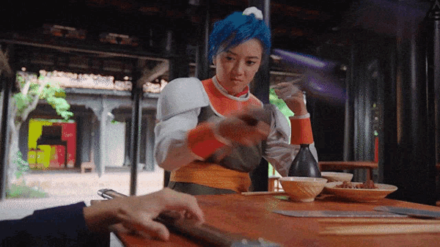 a woman with blue hair sits at a table with chopsticks and a sword