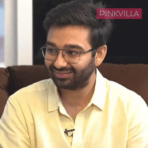 a man wearing glasses and a yellow shirt is smiling in front of a pinkvilla sign