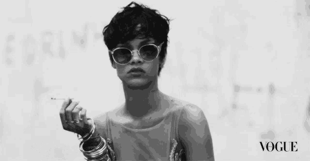 a black and white photo of a woman wearing sunglasses and holding a cigarette with vogue written on the bottom