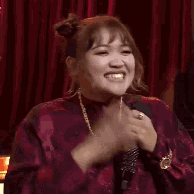 a woman wearing a purple dress and a gold chain is holding a microphone and making a funny face .