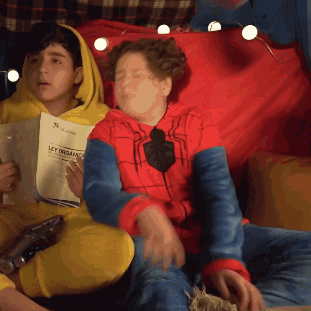 a boy in a spiderman costume sits next to a boy in a yellow hoodie reading a ley organica