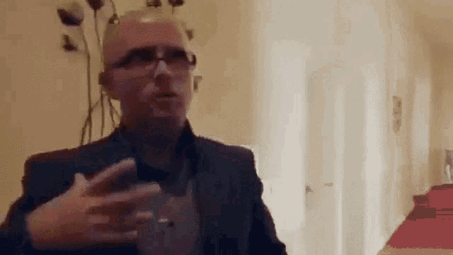 a bald man wearing glasses and a suit is talking to someone in a living room .