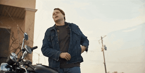 a man in a denim jacket is standing next to a black motorcycle