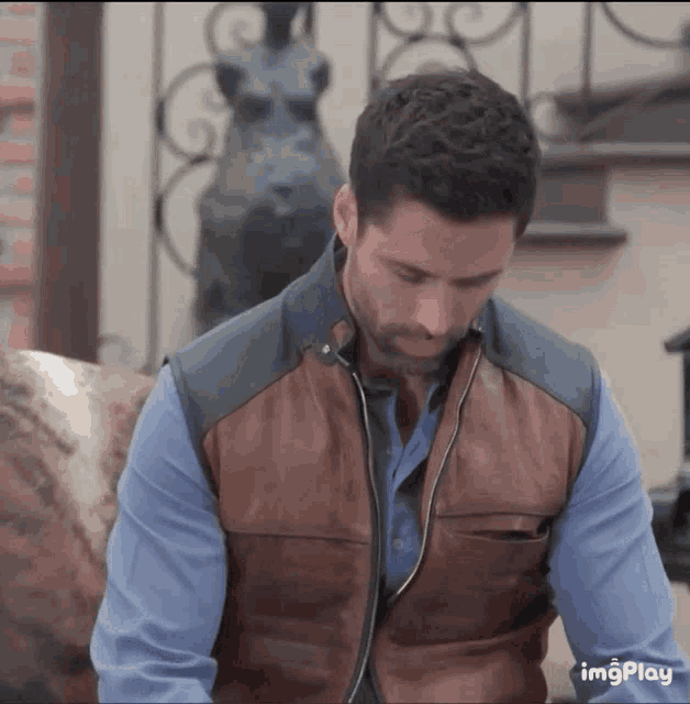 a man wearing a brown leather vest and a blue shirt is sitting in front of a statue