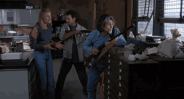 a man holding a shotgun stands next to a woman and another man in a messy room