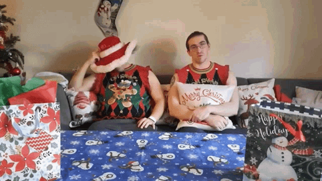two men wearing ugly christmas sweaters sit on a couch with a merry christmas pillow