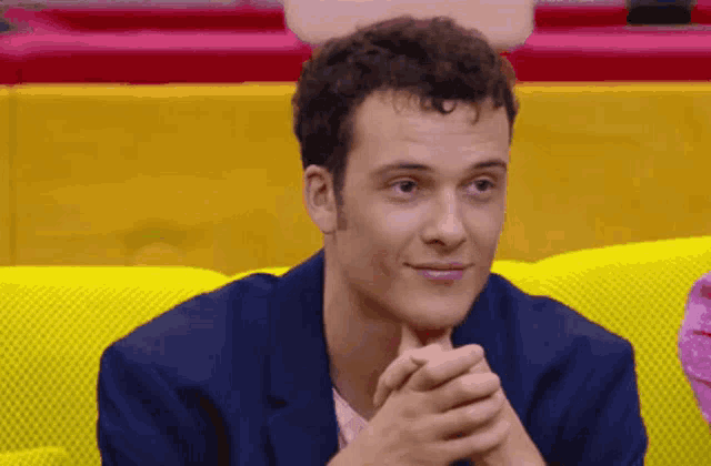 a young man in a blue suit is sitting on a yellow couch with his hands folded