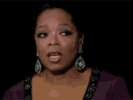 oprah winfrey is wearing a purple dress and earrings and is making a funny face .