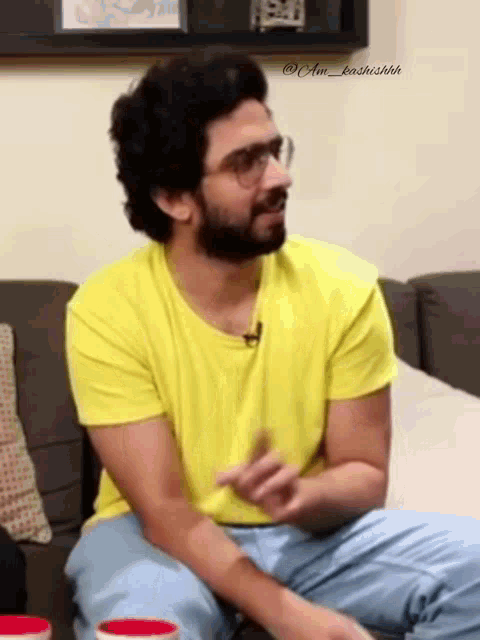 a man with a beard and glasses is sitting on a couch wearing a yellow shirt .