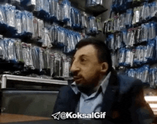 a man with a big nose is sitting at a table in front of a shelf full of cell phone cases .
