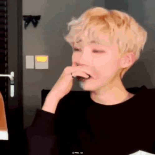 a young man with blonde hair is covering his mouth with his hand while wearing a black shirt .