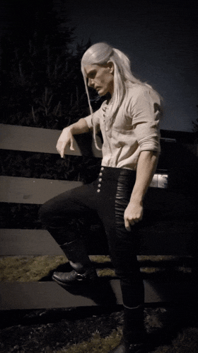 a man with long white hair leaning on a fence