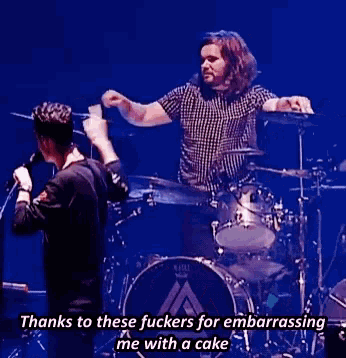a man in a plaid shirt is playing drums while another man sings into a microphone on stage .