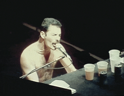 a shirtless man sings into a microphone while playing a piano