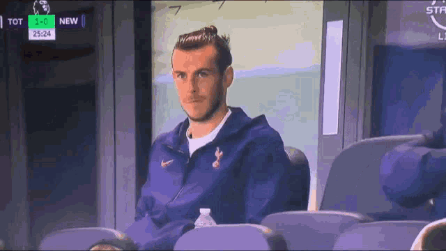 a man in a blue jacket is sitting in a stadium watching a soccer game between tot and new