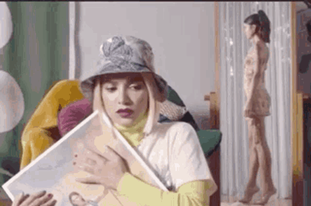 a woman in a bucket hat is holding a magazine while a woman in a dress is standing in the background .