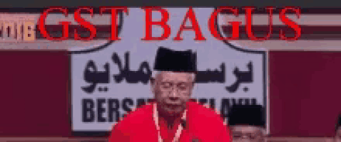 a man in a red shirt stands in front of a sign that says " gst bagus "