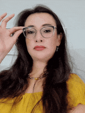 a woman wearing glasses and a yellow top holds her hand up