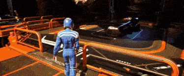 a man in a blue suit and helmet stands in front of a fence and a car