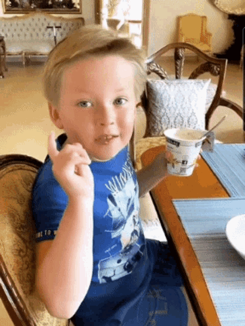 a young boy sitting at a table with a cup of yogurt in his hand