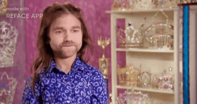 a man with long hair and a beard wearing a blue shirt is standing in front of a shelf full of crowns .
