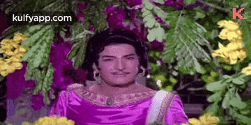 a man in a purple dress is standing in front of a bunch of flowers .