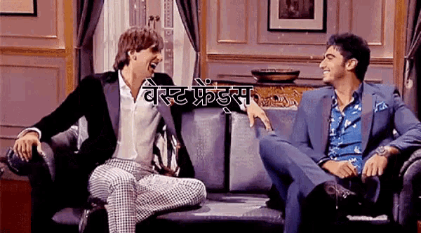 two men in suits are sitting on a couch and laughing in a room with a sign that says best friends on it