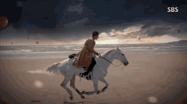 a man riding a white horse on the beach with sbs written on the bottom