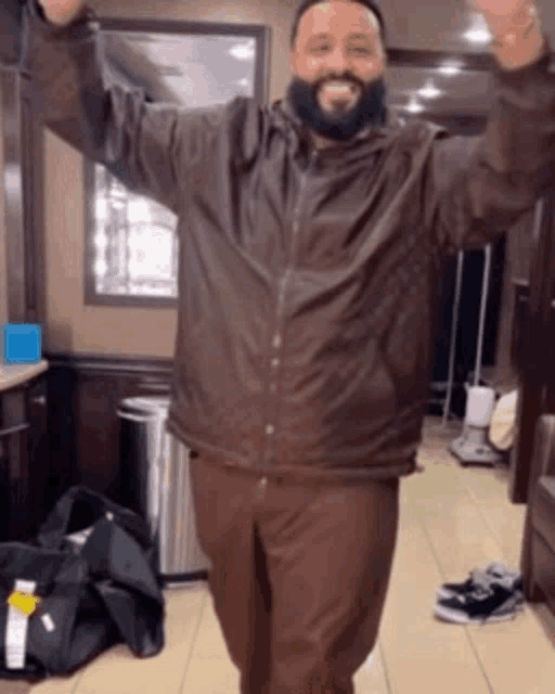 a man with a beard wearing a brown jacket is standing in a room with his arms in the air