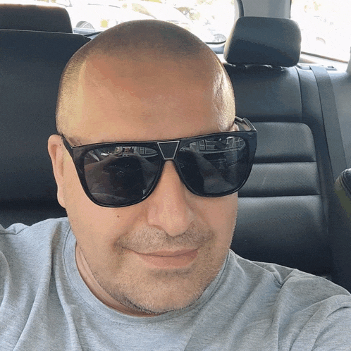 a man wearing sunglasses and a grey shirt smiles in a car