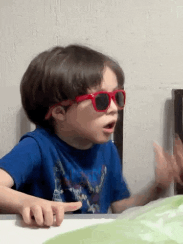a young boy wearing red sunglasses and a blue shirt is pointing at something .