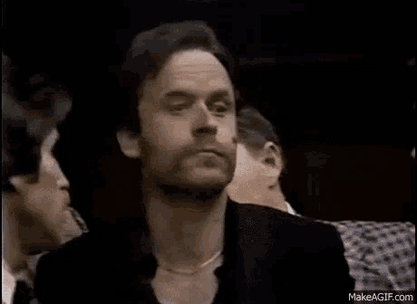 a man with a necklace around his neck is sitting in a room with other people .