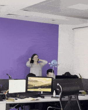 a woman adjusts her hair in front of a purple wall while sitting at a desk