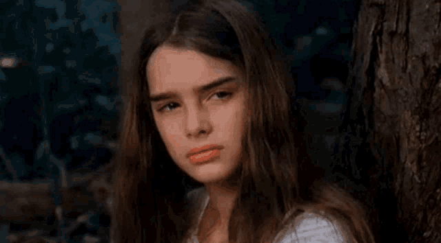 a young girl with long hair is leaning against a tree and making a sad face .