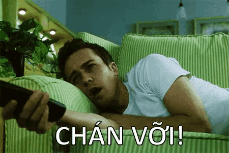 a man is laying on a green striped couch holding a remote control and saying chan voi !