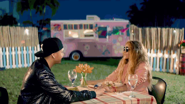 a man and a woman sitting at a table with a pink food truck in the background