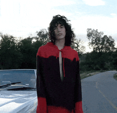 a man with curly hair wearing a red and black hoodie stands in front of a white car