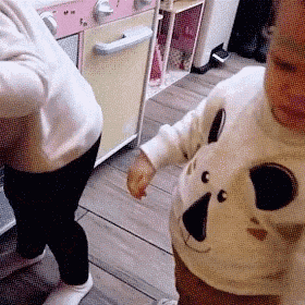 a baby wearing a white sweater with a panda face on it is standing next to a woman in a kitchen .