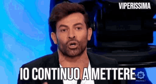 a man with a beard is giving a speech in front of a camera and says io continuo amettere .