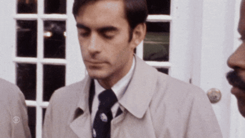 a man in a trench coat and tie is standing in front of a door