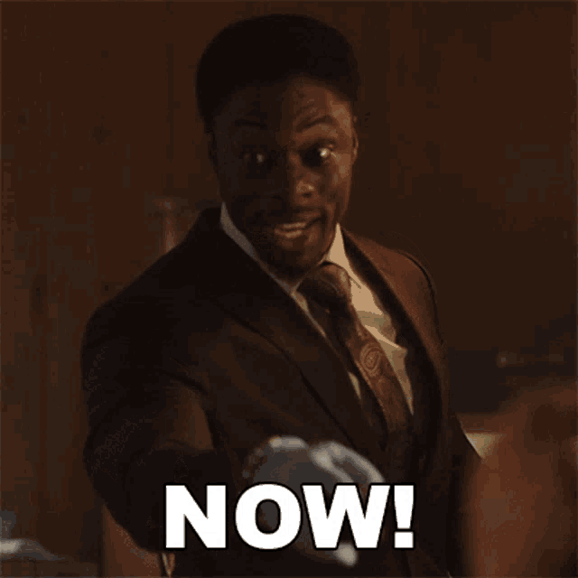 a man in a suit and tie is pointing at the camera with the word now written on his chest