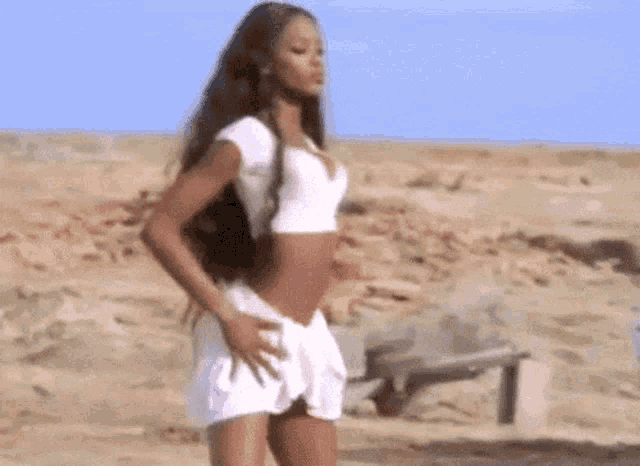 a woman in a white crop top and a white skirt is standing on a sandy beach .