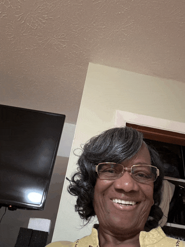 a woman wearing glasses and a yellow shirt is smiling in front of a flat screen tv